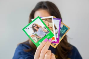 Young woman holding a fan of colorful printed photo cards, featuring branded frames from Spotify, Orange, Taco Bell, and scenic personal moments.