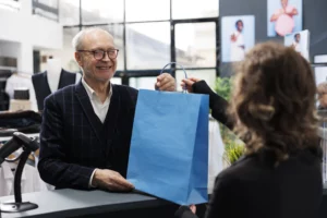 Elderly man smiling while receiving a blue shopping bag from a cashier at a modern retail clothing store counter.